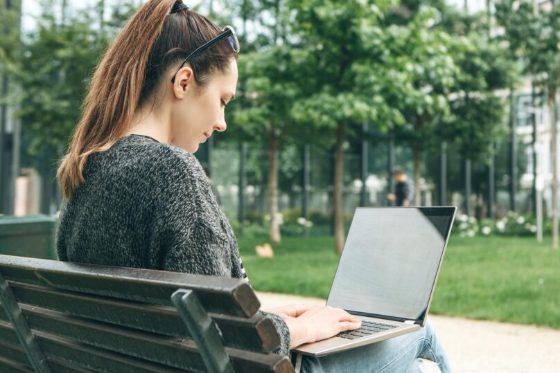 nainen istuu puiston penkilla ja kayttaa kannettavaa tietokonetta