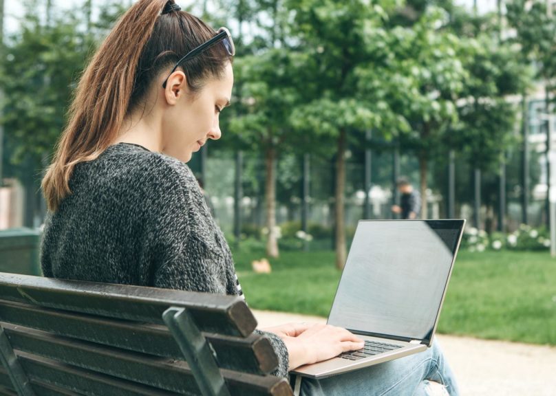nainen istuu puiston penkilla ja kayttaa kannettavaa tietokonetta