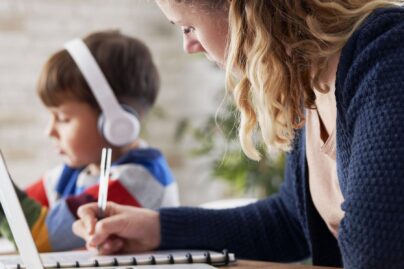 Äiti ja poika istuvat vierekkäin pöydän ääressä. Äiti tekee tietokoneella töitä ja poika on tabletilla kuulokkeet päässä.