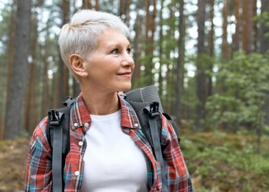 Harmaahiuksinen nainen kävelemässä metsässä, reppu selässä.