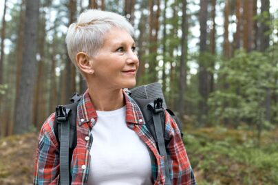 Harmaahiuksinen nainen kävelemässä metsässä, reppu selässä.