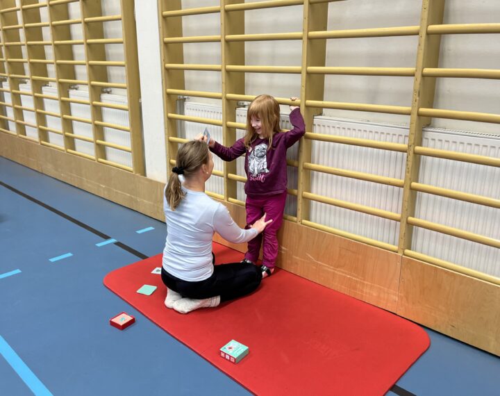 Aurora tekee seisaaltaan harjoitusta pidellen toisella kädellä puolapuusta kiinni. Fysioterapeutti Sari tukee jaloista.