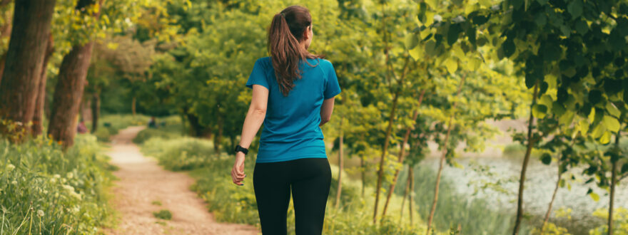 nainen kävelee kesäistä metsäpolkua vesistön lähellä