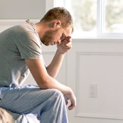 väsynyt mies nojaa käteensä sängyn reunalla