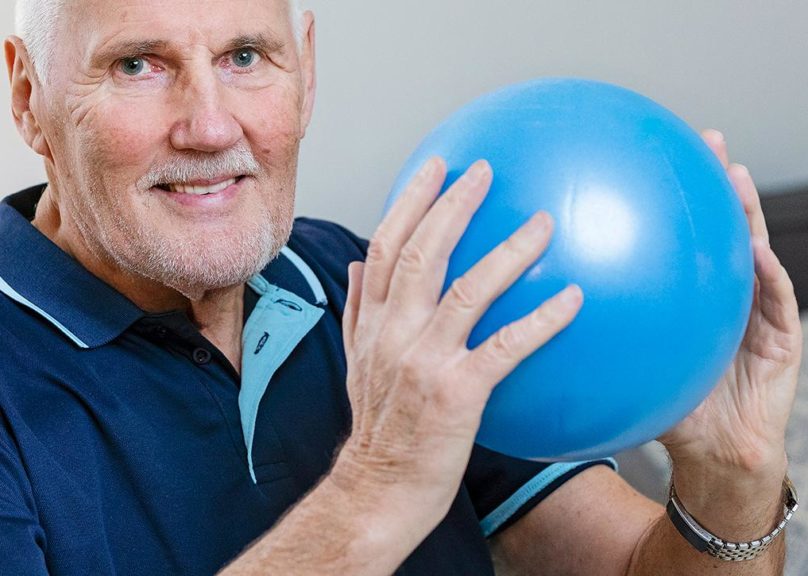 Seniorimies hymyilee ja pitää sinistä pientä jumppapalloa käsissään Coronarian kotifysioterapiassa.