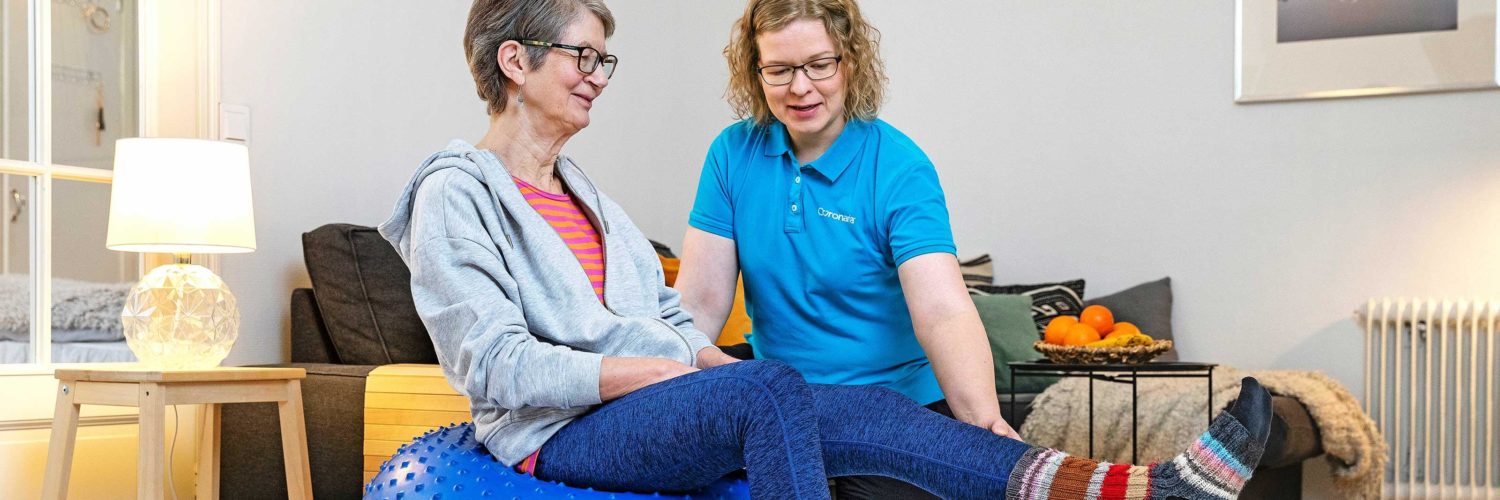 seniorinainen jumppaa sinisellä jumppapallolla ja naisfysioterapeutti avustaa sekä vieressä