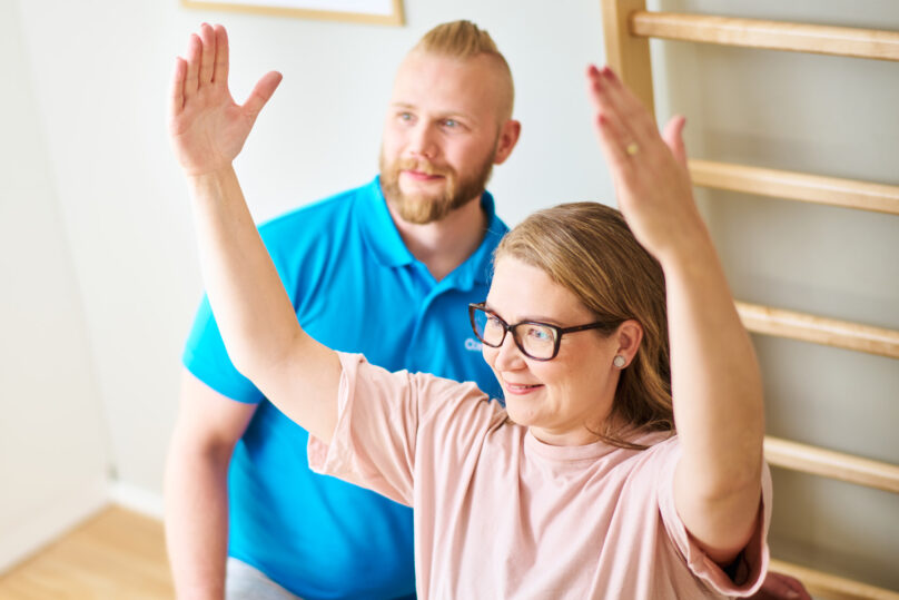 Naisasiakas tekee käsien nostoharjoitusta miesterapeutin ollessa taustalla tukena.