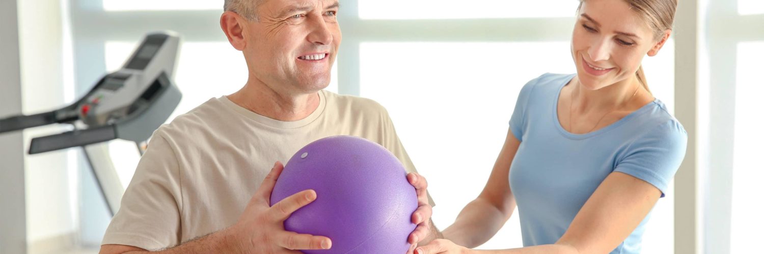 mies jumppaa pallon avulla ja fysioterapeutti ohjaa vieressa