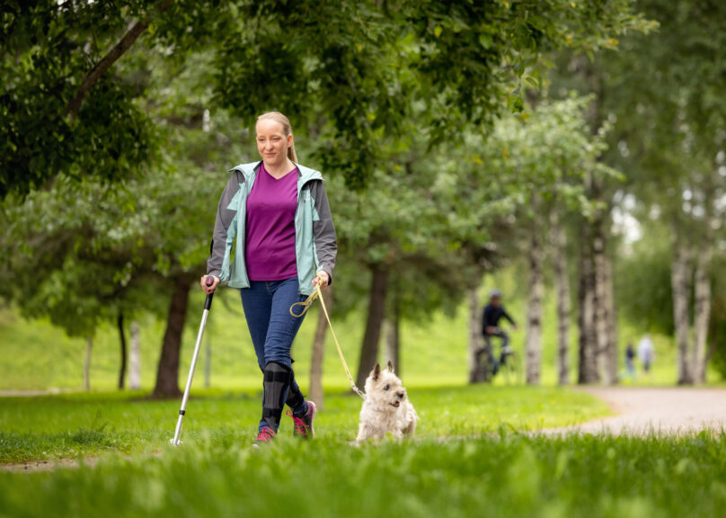 Vaaleahiuksinen nainen kävelee puistossa pienen koiran kanssa ja toisessa kädessä hänellä on kävelysauva.