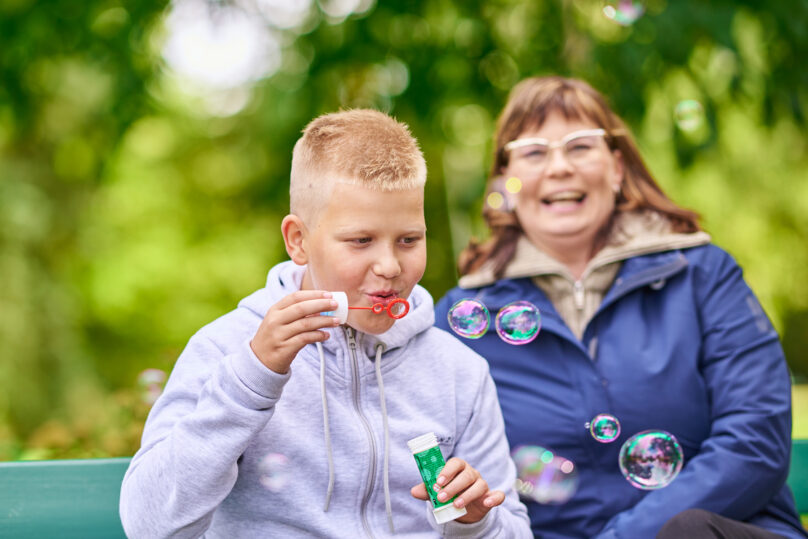 Lasten fysioterapiaan osallistuva poika puhaltaa saippuakuplia ja puistonpenkillä istuu hänen lähellään nainen.