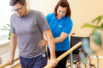 Naisterapeutti auttaa miesasiakasta neurologisen fysioterapian nojapuuharjoituksessa ja taustalla näkyy miehen pyörätuoli.