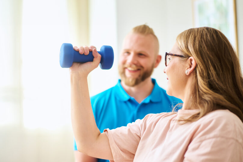 Naisasiakas tekee käsipainolla harjoitusta miesterapeutin ollessa taustalla tukena.