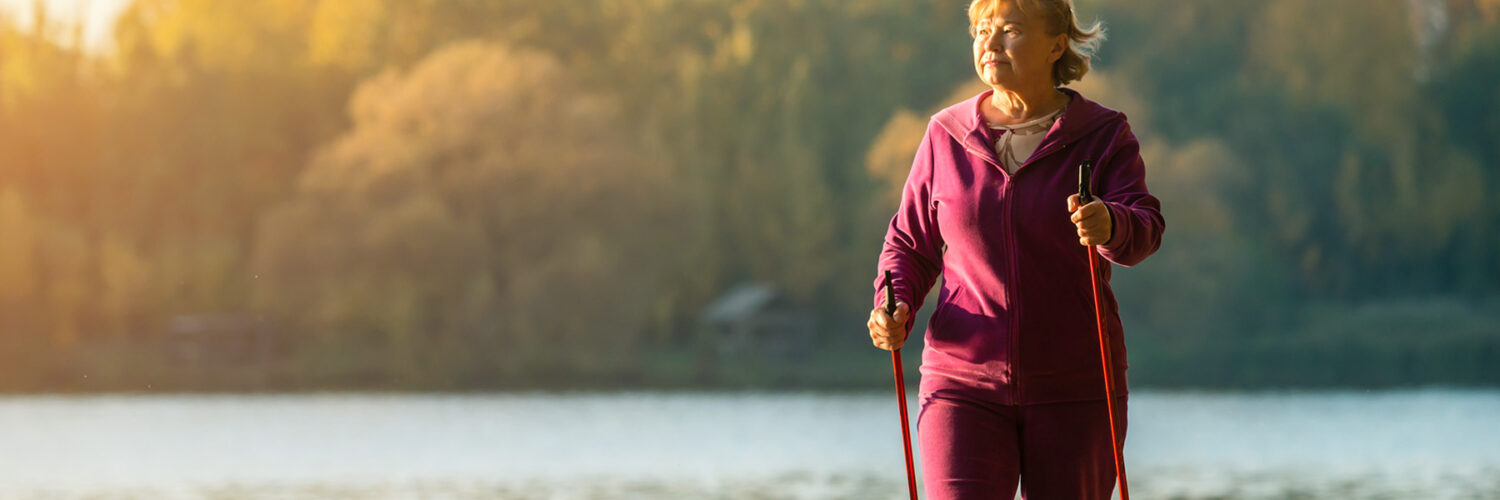 Iäkäs nainen sauvakävelee veden äärellä syysauringossa.