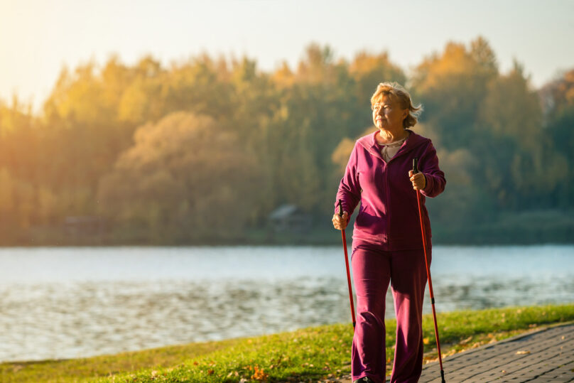 Iäkäs nainen sauvakävelee veden äärellä syysauringossa.