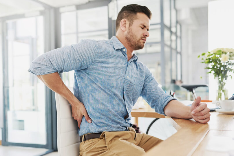 Tummahiuksinen mies istuu työpöydän ääressä ja pitää kättä alaselällä, joka on kipeä.