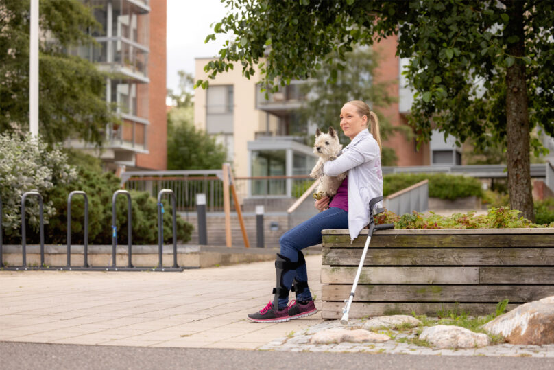 Vaaleahiuksinen nainen istuu istutusaltaan reunalla koira sylissään ja kävelysauva nojaa istutusallasta vasten.