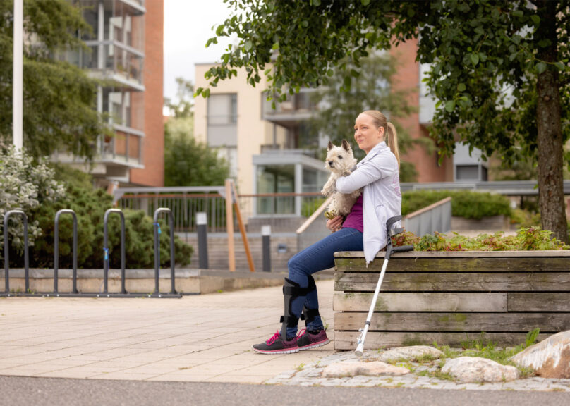 Vaaleahiuksinen nainen istuu istutusaltaan reunalla koira sylissään ja kävelysauva nojaa istutusallasta vasten.