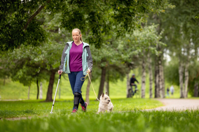 Vaaleahiuksinen nainen kävelee puistossa pienen koiran kanssa ja toisessa kädessä hänellä on kävelysauva.