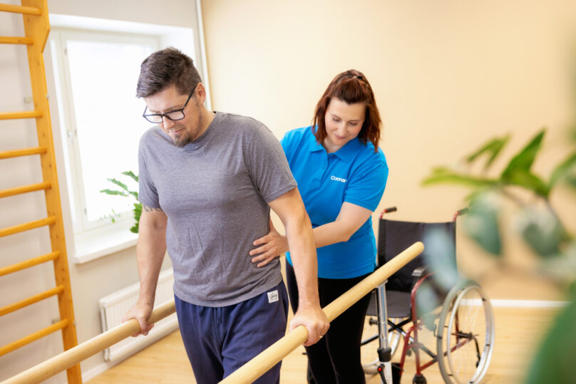 Naisterapeutti auttaa miesasiakasta nojapuuharjoituksessa ja taustalla näkyy miehen pyörätuoli.