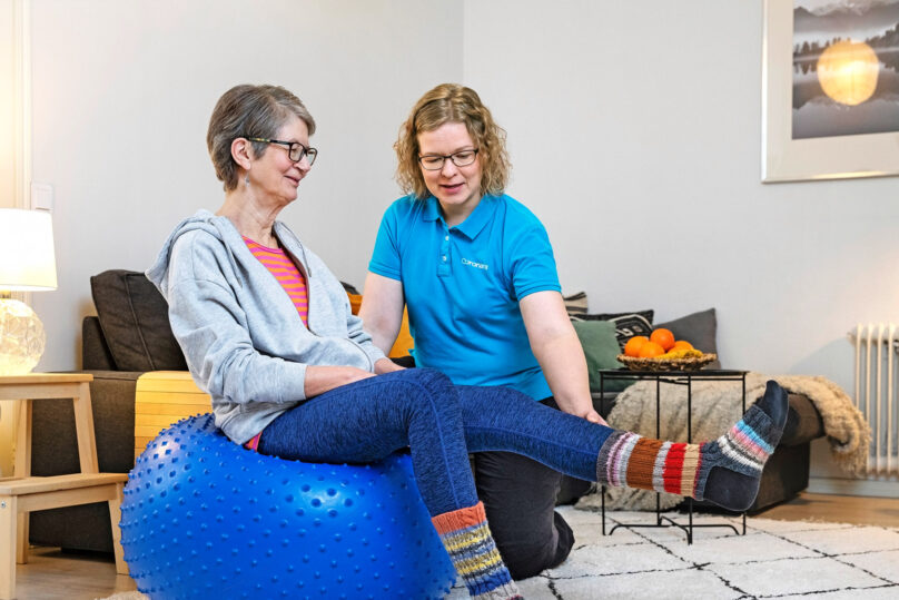 Iäkäs naisasiakas tekee jalan ojennusta jumppapallolla istuen ja naisterapeutti auttaa.