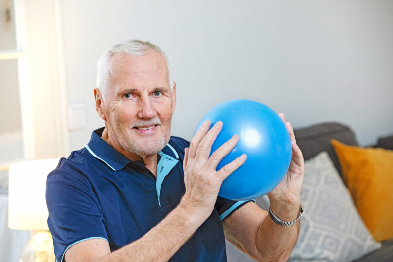 Seniorimies pitää sinistä jumppapalloa käsissä ja katsoo kohti.