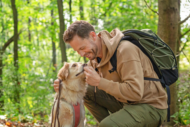 Tummahiuksinen mies koiran kanssa metsässä.