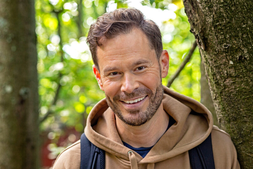 Tummahiuksinen mies on metsässä ja hymyilee lähikuvassa