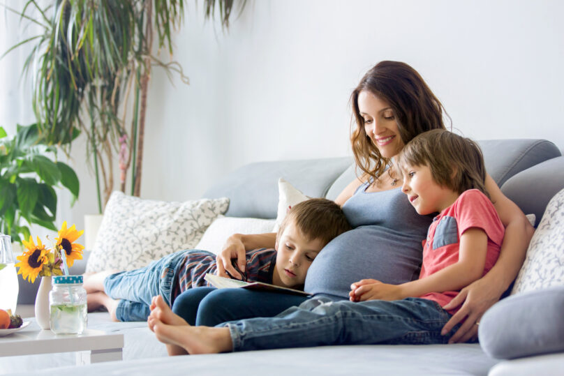Raskaana oleva nainen lukee sohvalla kirjaa kahden pojan kanssa.