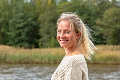 Vaaleahiuksinen nainen rannalla villapaidassa ja hymyilee kohti kameraa.