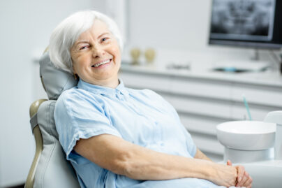 Seniorinainen, jolla hopeinen polkkatukka, istuu hoitotuolissa ja hymyilee.