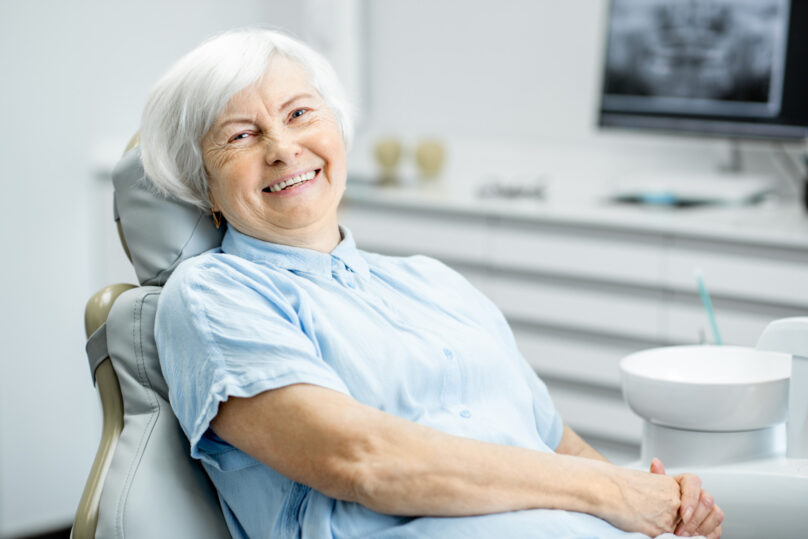 Seniorinainen, jolla hopeinen polkkatukka, istuu hoitotuolissa ja hymyilee.