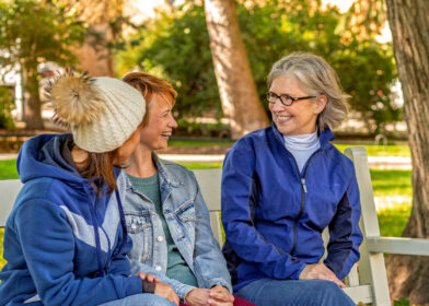 Kolme naista istuvat puiston penkillä ja hymyilevät.