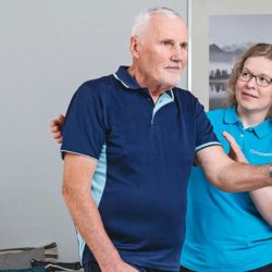 Seniorimiesasiakas keppijumppaa seisaaltaan ja fysioterapeutti ohjaa vieressä.