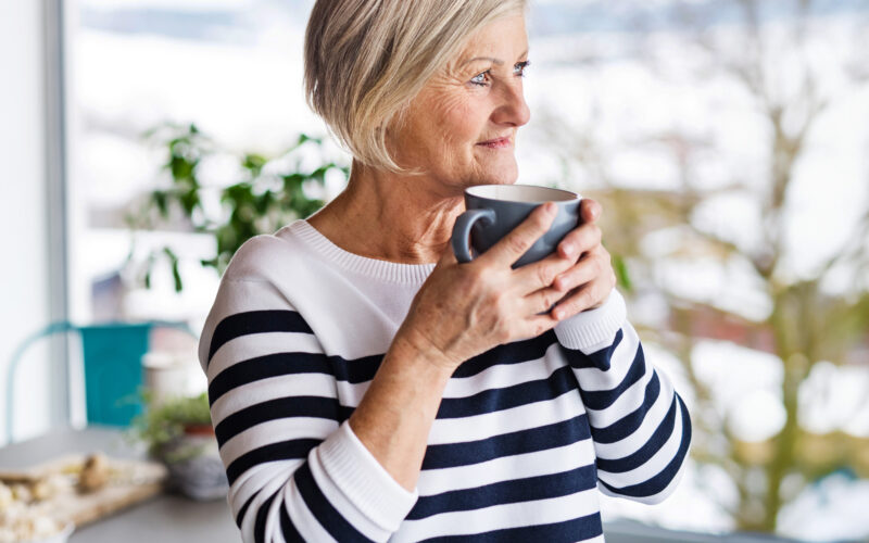 Iäkäs nainen kahvikuppi kädessä katsoo ulos.