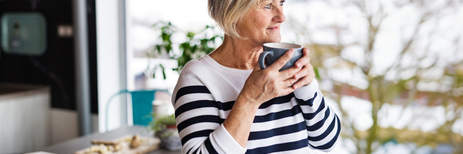 Iäkäs nainen kahvikuppi kädessä katsoo ulos.