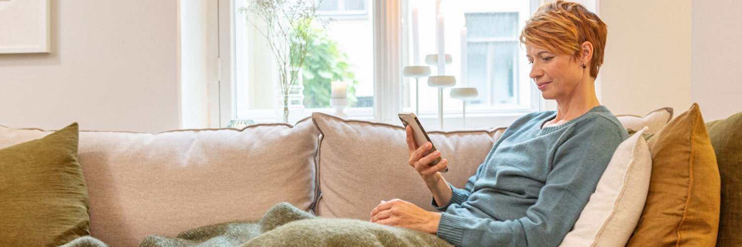 Punahiuksinen nainen istuu sohvalla ja katsoo puhelinta.