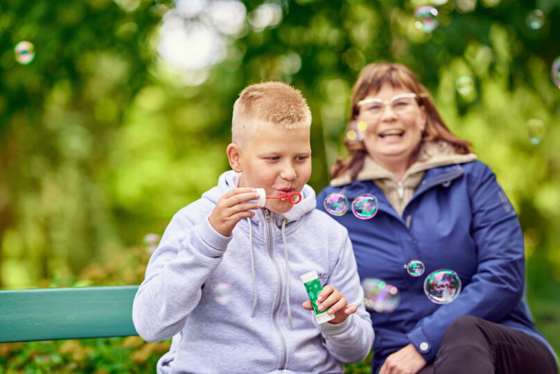 Äiti istuu puistonpenkillä ja poika puhaltaa vieressä saippuakuplia
