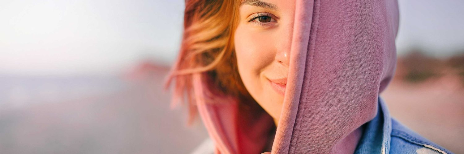 nuori nainen katsoo kameraan ja hymyilee auringonlaskussa rannalla huppu paassa