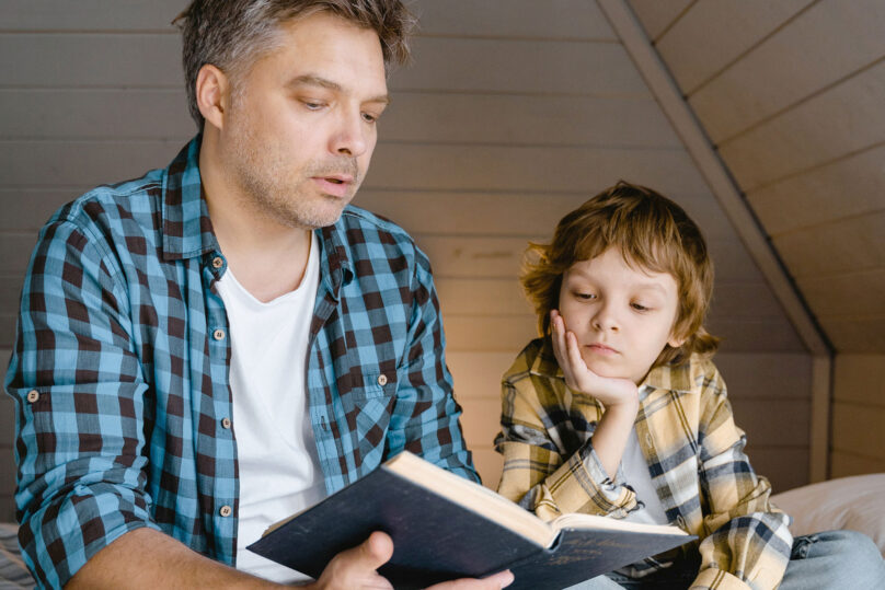 Lasten kehityksellisen kielihäiriön kuntoutukseen osallistuva poika ja isä istuu sängyllä ja lukee kirjaa..
