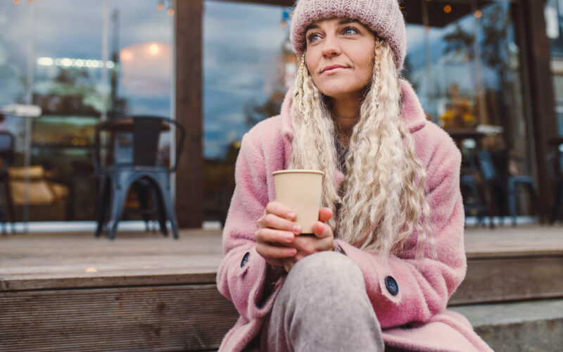 Vaaleahiuksinen nainen istuu ulkoterassin portailla pahvikuppi kädessä.