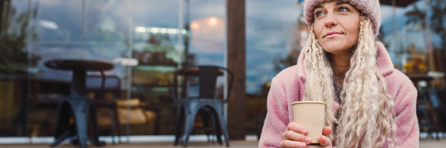 Vaaleahiuksinen nainen istuu ulkoterassin portailla pahvikuppi kädessä.