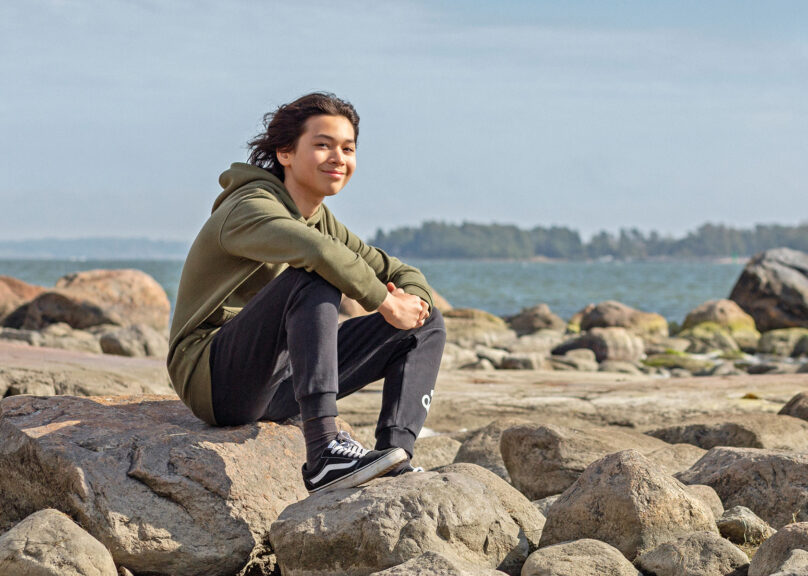 Poika ulkona hymyilee ja istuu rannalla kiven päällä