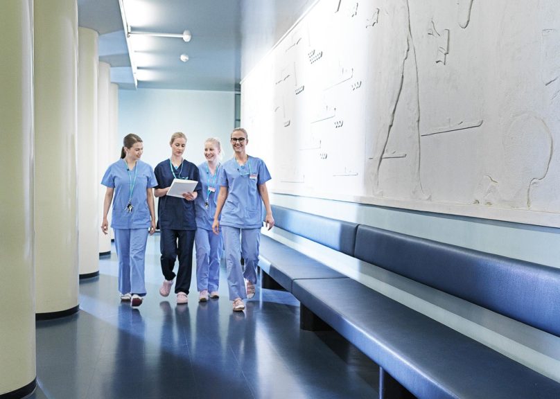 Sinisiin vaatteisiin pukeutuneet sairaanhoitajanaiset kävelevät käytävällä kohti kameraa.