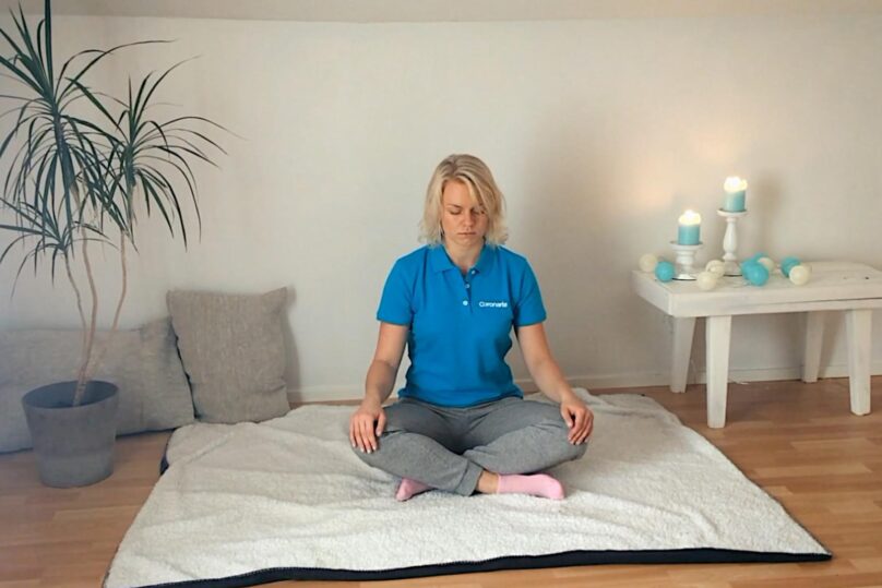 Sinipaitainen nainen istumassa lattialla jalat risti-istunnassa ja silmät kiinni.