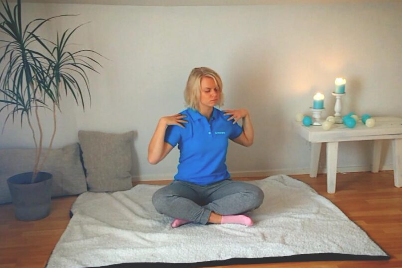 Sinipaitainen nainen istumassa lattialla jalat risti-istunnassa ja tekee ylävartalon pyöritysliikettä kädet harteilla.