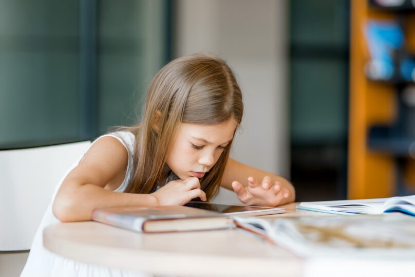 Ruskeahiuksinen tyttö on kumartunut lukemaan kirjaa kirjastossa.