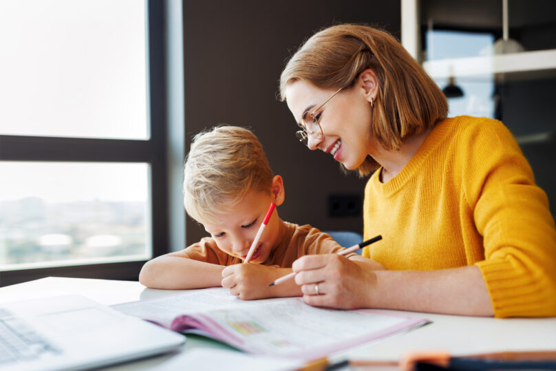 Vaaleahiuksinen poika ja äiti tekevät yhdessä läksyjä pöydän ääressä.