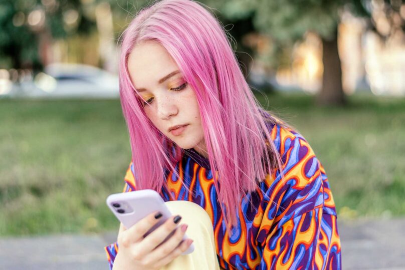 Pinkkihiuksinen nuori nainen katsoo kännykkää apeana puistossa.