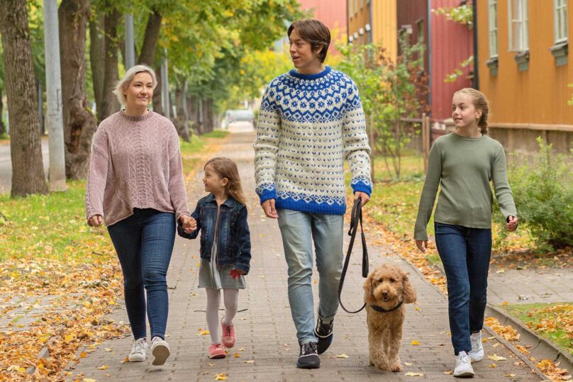 Äiti ja lapset kävelyttämässä koiraa kaupunkimaisemassa.