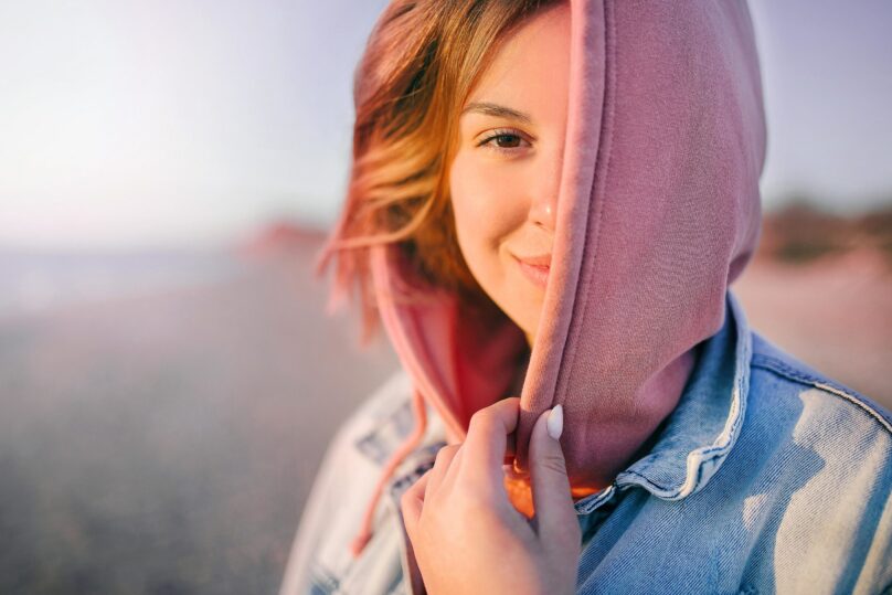 Nuori nainen katsoo kameraan hymyillen kasvot puoliksi vaaleanpunaisen hupun peitossa